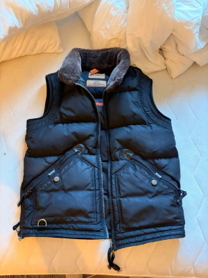 Parajumper väst - Kvitto finns! Snygg svart dunväst från Parajumpers med två stora dragkedjefickor framtill och detaljer i metall. Västen har en mjuk, grå fuskpäls runt kragen och orange detaljer bak. Perfekt för lager på lager och riktigt varm.