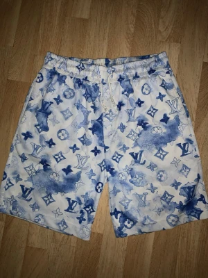 Blåvita Louis Vuitton shorts - Snygga shorts från Louis Vuitton med blått och vitt mönster och klassiska LV-loggor över hela tyget. De har elastisk midja med dragsko och är tillverkade i ett lätt material som passar perfekt till sommaren. Unik design som sticker ut.