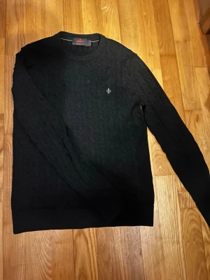 Svart kabelstickad tröja från Morris - Kabelstickad svart tröja från Morris med rund halsringning och diskret broderad logga på bröstet. Tröjan har lång ärm och är tillverkad i mjuk merinoull, perfekt för en stilren och klassisk look.