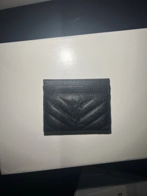 Svart kortfodral från Saint Laurent - Snyggt svart kortfodral från Saint Laurent i läder med diskret YSL-logga framtill. Fodralet har stilrena sömmar i chevronmönster och flera fack för kort. Perfekt accessoar för dig som gillar lyxiga detaljer och minimalistisk design.