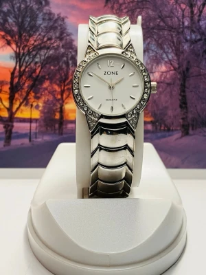 Silverfärgad klocka från Zone - Stilren klocka från Zone med silverfärgat armband i metall och rund vit urtavla. Urtavlan är dekorerad med gnistrande stenar runt kanten och har minimalistiska markeringar. Perfekt accessoar för att lyfta din outfit.Nytt batteri, säljes i befintligt skick enligt bilderna!