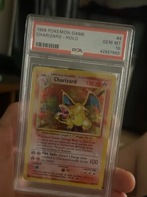 Charizard Pokémonkort 1999 PSA 10 - Charizard holografiskt samlarkort från 1999 Pokémon Game, graderat PSA 10 GEM MT. Kortet har en gul ram, färgstark illustration av Charizard och är inneslutet i en hård plastkapsel. Perfekt för Pokémonfans och samlare av trading cards.