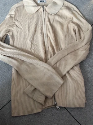 Beige ribbad ziptröja från JDY - Trendig beige ribbad tröja från JDY med dragkedja framtill och klassisk krage. Långärmad modell i mjukt stickat material som ger en snygg och avslappnad look. Perfekt att matcha med jeans eller kjol.