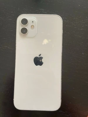Vit iPhone 12 med dubbla kameror - Snygg vit iPhone 12 med stilren design och två kameror på baksidan. Telefonen har en platt form med glasbaksida och aluminiumram. Apple-loggan sitter i mitten på baksidan och kameramodulen är placerad i det övre vänstra hörnet.