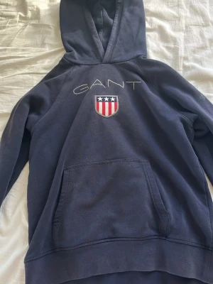 Marinblå hoodie från GANT - Snygg marinblå hoodie från GANT med broderad logga och sköld på bröstet. Klassisk känguruficka framtill och stor huva. Perfekt för en avslappnad och sportig stil. Mjuk bomullsblandning som känns skön mot huden. (den har ett hål på magfickan, som inte syns när man har den på sig)