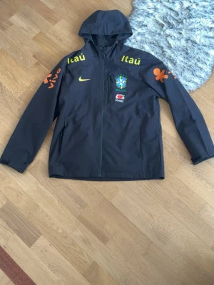 Svart Brasil Nike Vindjacka  - Svart vindjacka från Nike med huva och dragkedja. Jackan har Brasilien landslagets emblem, gula och orange tryck med sponsorer samt logga på bröstet. Perfekt för fotbollsälskare!!                                                (Jackan är i Storlek L men passar bättre för dig som är Storlek M!)