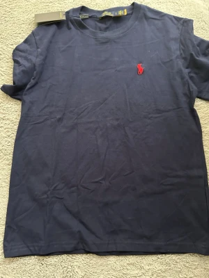 Marinblå t-shirt från Polo Ralph Lauren - Snygg marinblå t-shirt från Polo Ralph Lauren med klassisk röd broderad logga på bröstet. Tillverkad i mjuk bomull och har en normal passform med korta ärmar och rund hals. Perfekt basplagg för en clean och stilren look.