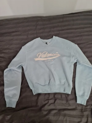 Ljusblå sweatshirt Valencia Spain - Ljusblå sweatshirt från Diveded med broderad text 'Valencia Spain' i vitt på bröstet. Tröjan har rund halsringning, ribbade muddar vid ärmslut och nederkant samt en avslappnad passform. Perfekt för en chill och stilren look.