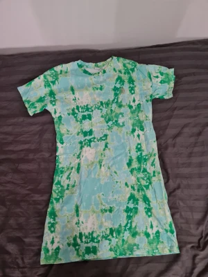 Grön och blå tie-dye t-shirtklänning - Cool t-shirtklänning med tie-dye mönster i grönt, blått och vitt. Klänningen har korta ärmar och rak passform, perfekt för en avslappnad stil. Tillverkad i mjukt bomullsmaterial som känns skönt mot huden.