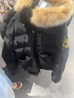 Svart dunjacka från Zavetti Canada - Snygg svart dunjacka från Zavetti Canada med stor huva och fluffig fuskpäls. Jackan har två stora fickor framtill, dragkedja och tryckknappar samt tydliga logopatchar på ärmen och bröstet. Perfekt för kalla vinterdagar.
