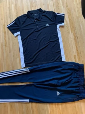 Adidas träningsset svart/vit - Snyggt träningsset från Adidas med svart pikétröja med vita detaljer och matchande svarta träningsbyxor med vita ränder längs sidorna. Båda plaggen har Adidas-logga och är tillverkade i ett lätt och bekvämt material, perfekt för sport och träning.