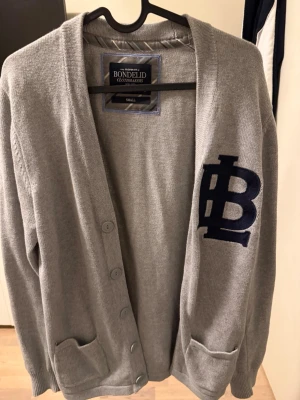 Grå kofta från Bondelid - Grå kofta från Bondelid med mörkblå broderat emblem på bröstet, knäppning framtill och två fickor. Mjukt stickat material och svarta mockadetaljer på armbågarna. Perfekt för en chill och stilren look.