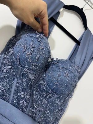 Lång blå klänning med spetsdetaljer - Elegant långklänning i blått med vackra spetsdetaljer och blommigt mönster på överdelen. Klänningen har vadderad byst, markerad midja och draperad kjol. Perfekt för bal eller festliga tillfällen.