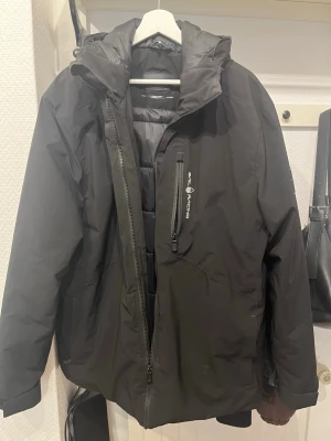 Svart pufferjacka från Sail Racing - Svart pufferjacka från Sail Racing med hög krage, dragkedja framtill och bröstficka med logga. Jackan har vadderad insida och en stilren, sportig look. Perfekt för kalla dagar när du vill hålla värmen och se snygg ut.
