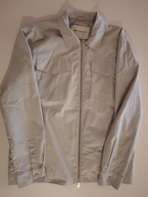 Ljusgrå overshirt från These Glory Days XL (Som Large) - Stilren ljusgrå overshirt med dragkedja framtill från These Glory days. Modellen har klassisk krage, två bröstfickor med lock och knäppning vid ärmslut. Tillverkad i ett lätt och mjukt bomullstyg, perfekt för lager-på-lager.