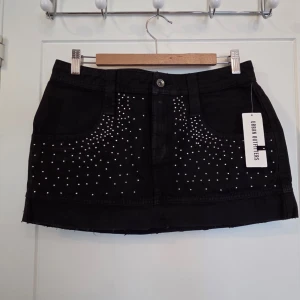 Svart kortkjol med nitar Urban Outfitters - Cool svart kortkjol från Urban Outfitters med silvriga nitar framtill. Kjolen har klassisk jeansmodell med fickor och bälteshällor. Perfekt för dig som gillar en edgy stil och vill sticka ut lite extra.