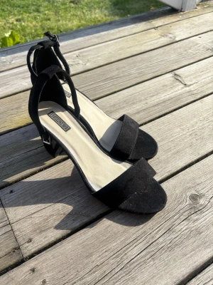 Svarta klackpumps från Bianco - Stilrena svarta pumps från Bianco med öppen tå, bred klack och justerbart ankelband med silverfärgat spänne. Skorna har en mjuk innersula och ovandel i mockaimitation som ger en lyxig känsla. Perfekta för dig som vill ha både stil och komfort.