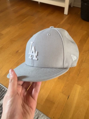 Ljusgrå LA-keps från New Era - Snygg ljusgrå keps från New Era med LA-logga broderad framtill i vitt. Klassisk modell med böjd skärm och broderade ventilationshål. Kepsen är helfärgad och har även New Era-loggan på sidan och MLB-logga bak. Perfekt för dig som gillar streetwear och sportig stil.