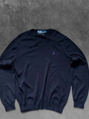 Svart v-ringad tröja från Polo Ralph Lauren - Svart v-ringad tröja från Polo Ralph Lauren med lila broderad logga på bröstet. Tröjan har långa ärmar och ribbade muddar vid ärmslut och nederkant. Perfekt för en stilren och klassisk look.