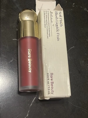 Rare Beauty Soft Pinch Liquid Blush - Flytande rouge från Rare Beauty i nyansen 'Believe', en djup rosabrun färg. Kommer i en frostad glasflaska med guldig rund kork och elegant design. Perfekt för att skapa en naturlig och fräsch look med enkel applicering.