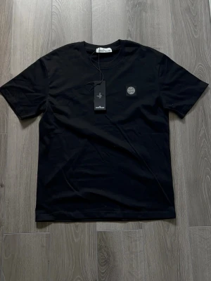 Svart t-shirt från Stone Island - Snygg svart t-shirt från Stone Island med klassisk logotyp på bröstet. T-shirten har rund halsringning och korta ärmar. Tillverkad i mjuk bomull som känns skön mot huden. Perfekt för en clean och stilren look.
