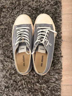 Grå sneakers från arigato  - Snygga ljusblå sneakers från arigato med klassisk låg design. Skorna har ovandel i canvas, vita gummisulor och tåhätta samt matchande grå skosnören. Perfekta för en stilren och avslappnad look.