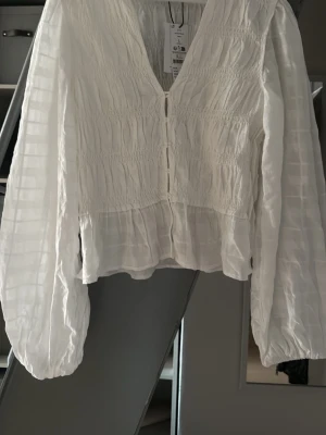 Vit smockad blus med v-ringning - Vit oanvänd blus från Ginatricot med smockdetaljer och v-ringning. Blusen har långa, luftiga ärmar och knäppning framtill. 