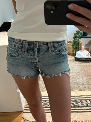 Blå jeansshorts med fransig kant - Snygga blå jeansshorts med fransig, rå kant och klassisk femficksdesign. Shortsen har bälteshällor och knappgylf. Perfekta för varma dagar och en avslappnad stil.