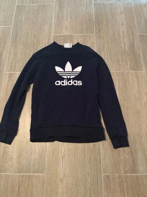 Mörkblå Adidas sweatshirt - Snygg mörkblå sweatshirt från Adidas med det klassiska vita Trefoil-loggan tryckt på bröstet. Tröjan har rund hals, långa ärmar och ribbade muddar. Perfekt för en sportig och avslappnad stil.
