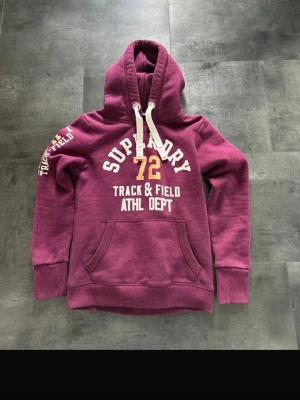 Vinröd hoodie från Superdry - Snygg vinröd hoodie från Superdry med vit och gul text på bröstet och ärmen. Klassisk känguruficka framtill, vit dragsko i huvan och mjukt material. Perfekt för en sportig och avslappnad stil.