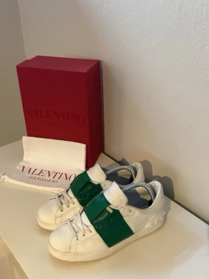 Valentino sneakers med allt og - Nästan helt brand new Snygga vita sneakers från Valentino Garavani med en bred grön läderdetalj över mitten. Skorna har klassisk låg modell, vita snören och subtila nitar på hälen. Perfekta för dig som vill sticka ut med en lyxig touch. 