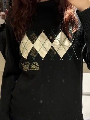Svart argyletröja från Ecko Unltd - Snygg svart stickad tröja från Ecko Unltd med klassiskt argylemönster i vitt, beige och grönt framtill. Broderad logga i gult nedtill. Tröjan har rund halsringning och är perfekt för dig som gillar streetwear med en twist.