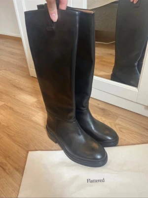 Svarta knähöga läderstövlar Flattered - Säljer ett par svarta knähöga stövlar från Flattered i slätt läder. Klassisk rund tå, robust sula och minimalistisk design. Perfekta för dig som vill ha en stilren och tidlös look till hösten och vintern.