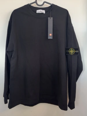 Stone Island tröja - Svart långärmad tröja från Stone Island med klassisk rund hals och ikonisk logotyp-patch på vänster ärm. Tröjan är i mjuk bomull och har en avslappnad passform. Perfekt för en stilren och trendig look.