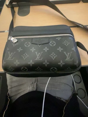 Svart axelväska  - Snygg svart axelväska med klassiskt monogrammönster i grått. Väskan har en silverfärgad dragkedja och justerbar axelrem. Perfekt för att bära dina viktigaste saker med stil.