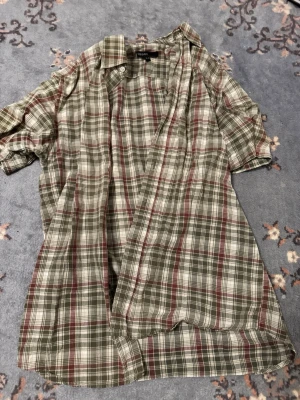 Grönrutig kortärmad skjorta - Kortärmad skjorta från Woodland i grön, vit och röd rutig design. Skjortan har klassisk krage, knappar framtill och är tillverkad i lätt bomullsmaterial. Perfekt för en avslappnad och cool stil.