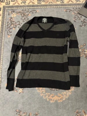 Randig stickad tröja med V-ringning - Snygg stickad tröja från WeSC med breda svarta och mörkgrå ränder. Tröjan har V-ringning, långa ärmar och ribbade muddar vid ärmslut och nederkant. Perfekt för en avslappnad och stilren look.