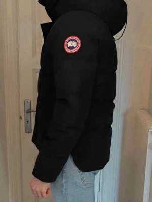 Svart dunjacka från Canada Goose - NYPRIS - 13.5699.                                    Säljer en svart dunjacka från Canada Goose med klassisk patch på ärmen och huva. Jackan är vadderad och har en stilren, rak passform. Perfekt för kalla vinterdagar och håller dig varm med sin rejäla dunfyllning. 
