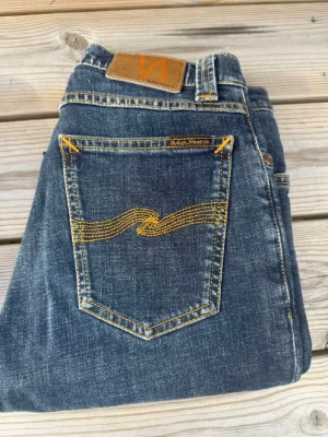Mörkblå jeans från Nudie Jeans - Snygga mörkblå jeans från Nudie Jeans med klassisk femficksdesign och gul kontrastsöm på bakfickorna. Jeansen har rak passform och är tillverkade i slitstark denim. Perfekta för en avslappnad och trendig look. High kai modell