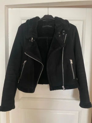 Svart pälsjacka med dragkedjor - Snygg svart pälsjacka från ZARA med silvriga dragkedjor och mjukt foder. Jackan har en klassisk bikerstil med bred krage och detaljerade metalldragkedjor på fickor och framsida. Perfekt för kalla dagar och ger en cool look.