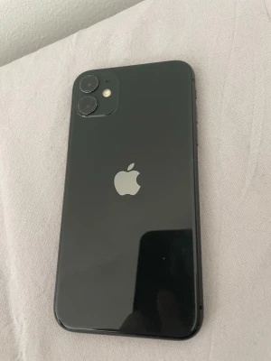  iPhone 11 med dubbla kameror - Snygg iPhone 11 med stilren design och rundade kanter. Baksidan är i glas och har två kameror samt blixt i hörnet. Apple-loggan sitter i mitten. Perfekt för dig som vill ha en modern och kraftfull smartphone.