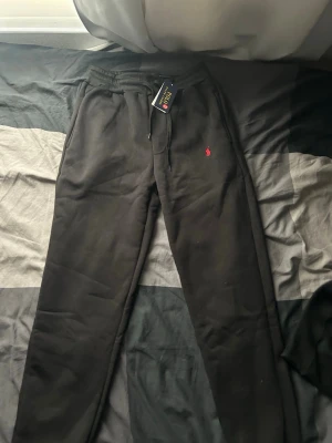 Svarta mjukisbyxor från Polo Ralph Lauren - Svarta mjukisbyxor från Polo Ralph Lauren med dragsko i midjan och ribbade muddar vid bensluten. Klassisk röd logga broderad på vänster sida. Byxorna har sidofickor och är tillverkade i mjukt material för maximal komfort.