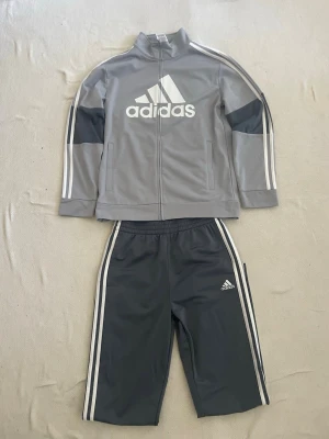  Adidas tracksuit - Snyggt träningsset från Adidas i två nyanser av grått med vita detaljer. Jackan har dragkedja, ståkrage och stora Adidas-loggan framtill. Byxorna har resår i midjan och klassiska vita ränder längs sidorna samt liten logga på benet. Perfekt för sport eller chill. Barn storlek L, 14-16.