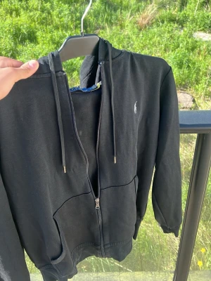 Svart hoodie från Ralph Lauren - Svart hoodie med dragkedja från Ralph Lauren. Klassisk design med huva och dragsnören, två fickor framtill och den ikoniska vita loggan broderad på bröstet. Perfekt för en chill och stilren look.