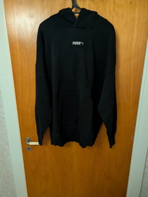 Svart hoodie från Puma - Svart hoodie från Puma med klassisk logga på bröstet. Hoodien har en stor känguruficka framtill och en rymlig huva. Perfekt för en avslappnad och sportig stil. Tillverkad i mjukt material som känns skönt mot huden.