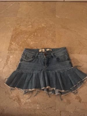 Blå volang jeanskjol med rå kant - Säljer en blå jeanskjol med volang och råa kanter. Kjolen har knapp- och dragkedjestängning samt en kort, utställd modell. Perfekt för dig som gillar en edgy och trendig stil. Egensydd av ett par gamla denim jeans från Sween. 