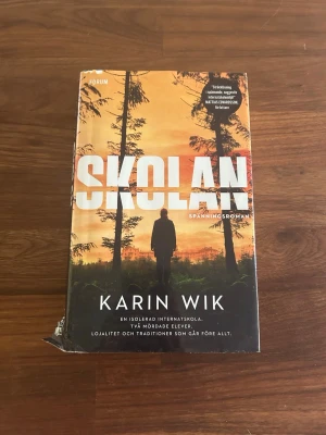 Skolan av Karin Wik - Spänningsroman 'Skolan' av Karin Wik. Omslaget har en siluett av en person i en skog med orange och svarta toner. Boken är inbunden och har ett dramatiskt och mystiskt tema kring en isolerad internatskola och mordgåtor.