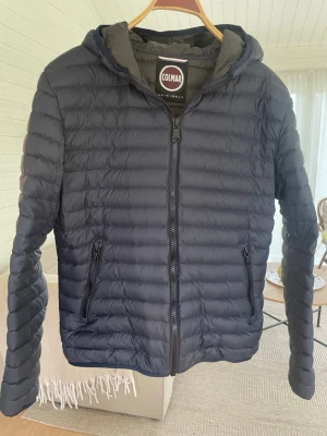 Mörkblå dunjacka från Colmar - Snygg mörkblå dunjacka från Colmar med huva och dragkedja framtill. Jackan har två fickor med dragkedja och quiltad design. Perfekt för kyliga dagar och enkel att matcha med olika outfits. Jackan är för liten för mig och därför tyvär inte kommit till andvändning och bara hängt i garderoben