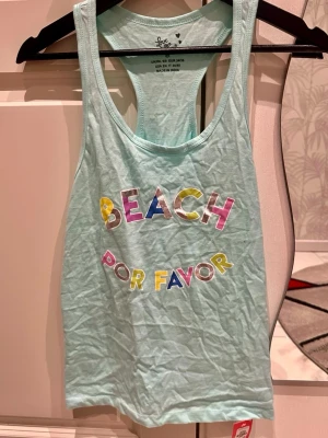 Mintgrön linnetopp med färgglatt tryck - Snygg mintgrön linnetopp med texten 'BEACH POR FAVOR' i färgglada bokstäver framtill. Modellen är ärmlös med brottarrygg och tillverkad i mjuk bomull, perfekt för varma dagar.