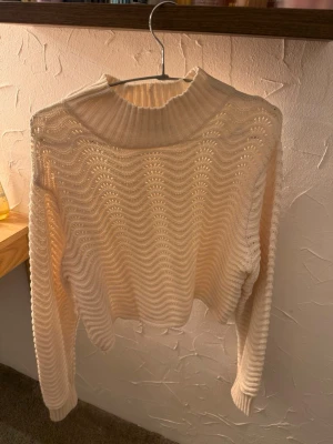 Beige stickad tröja med vågmönster - Trendig beige stickad tröja med vågigt mönster och hög ribbad krage. Tröjan har långa ärmar och är tillverkad i ett mjukt, tunt material som ger en luftig känsla. Perfekt för lager-på-lager och snygg till både jeans och kjol.